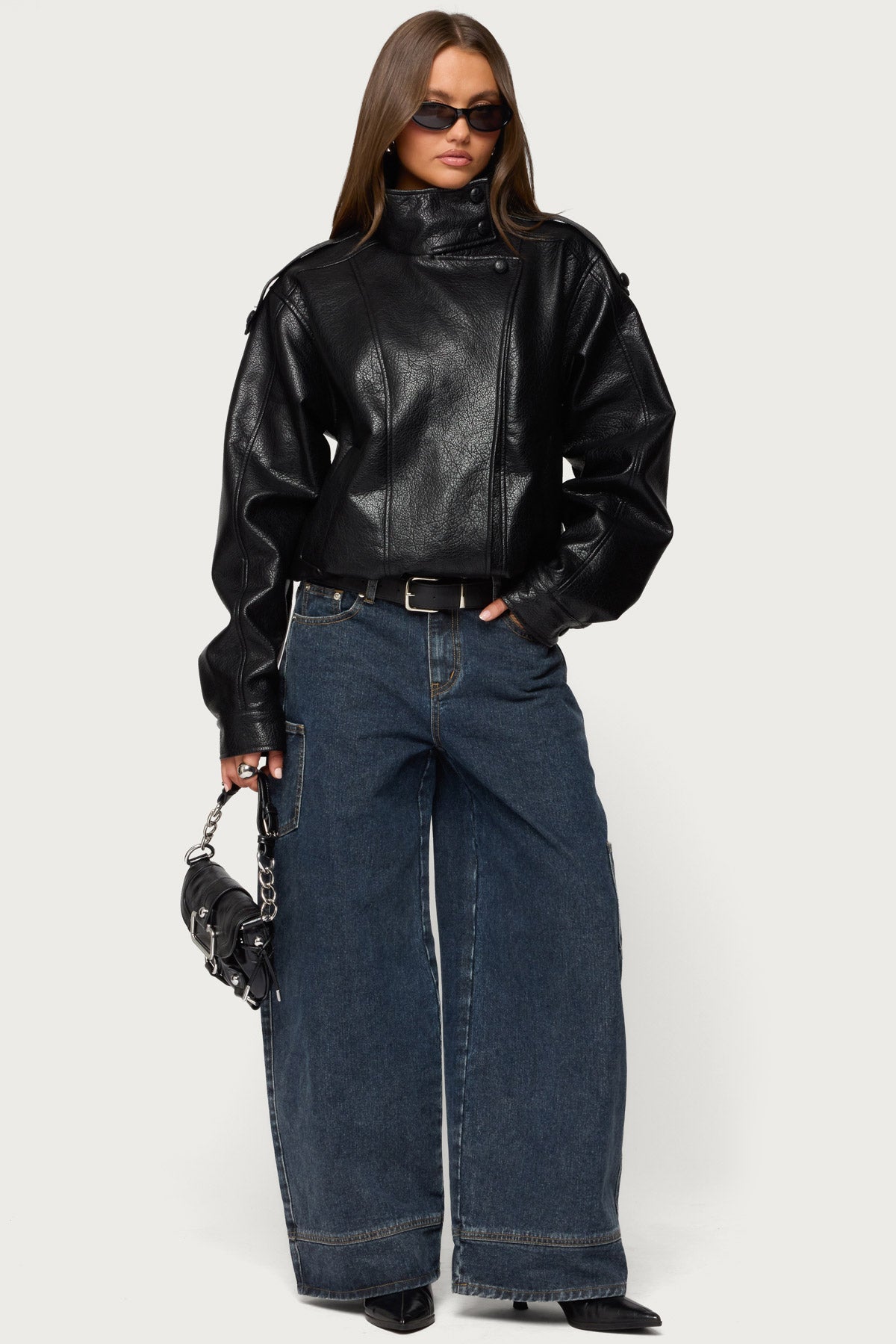 Ganzkörperbild einer Frau mit Sonnenbrille in schwarzer Oversize-Kunstlederjacke, weiter dunkler Cargo-Jeans, Gürtel und kleiner schwarzer Tasche mit Kette